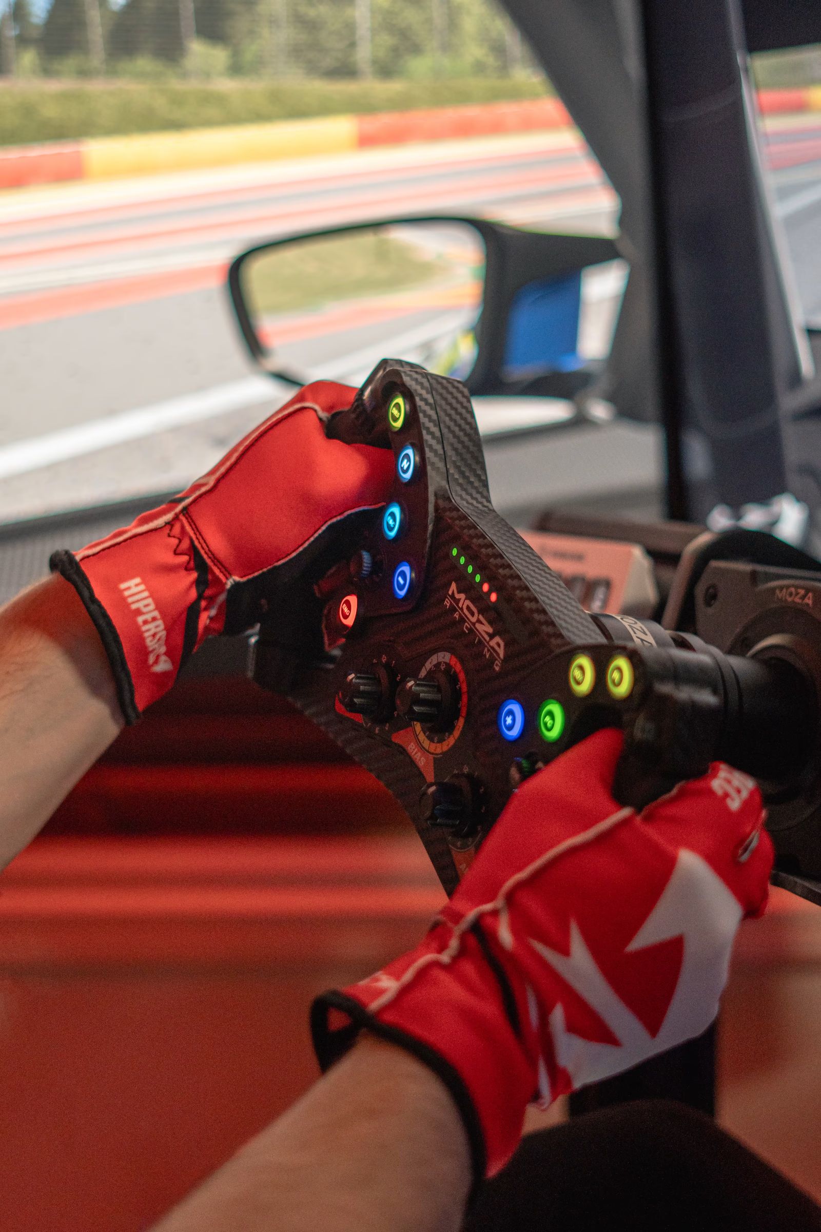 Hands in Crimson gloves gripping a sim racing wheel, the track on screen behind.